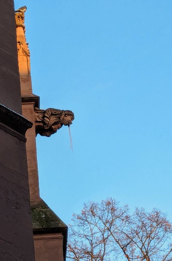 Statue, die mit dem Gesicht nach unten an einer Kirche hängt. Aus ihrem Mund kommt ein großer Eiszapfen, es sieht aus als ob die Statue von oben runterkotzt.
