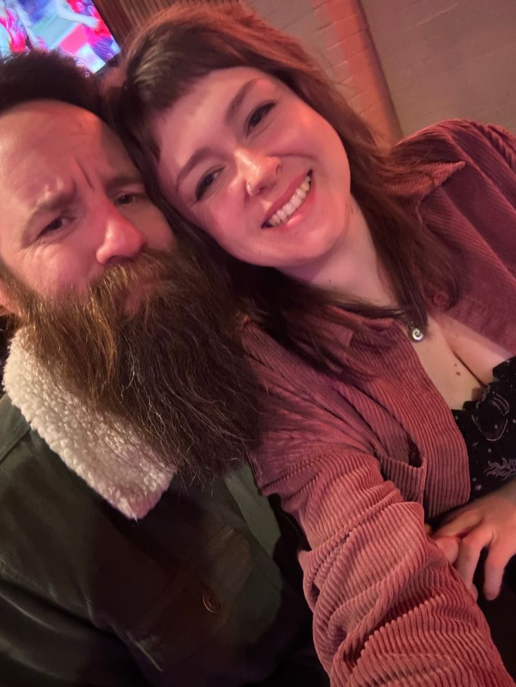 mum (me) and dad (steven) smiling at a bar. my boobies are out a bit. there’s footy on the tv in the background (spew)