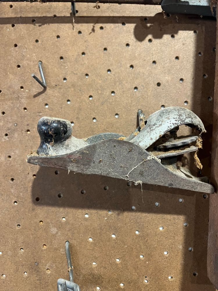 Another small metal antique hand plane hanging on a pegboard, this one caked in cobweb-covered sawdust and pitted with rust.