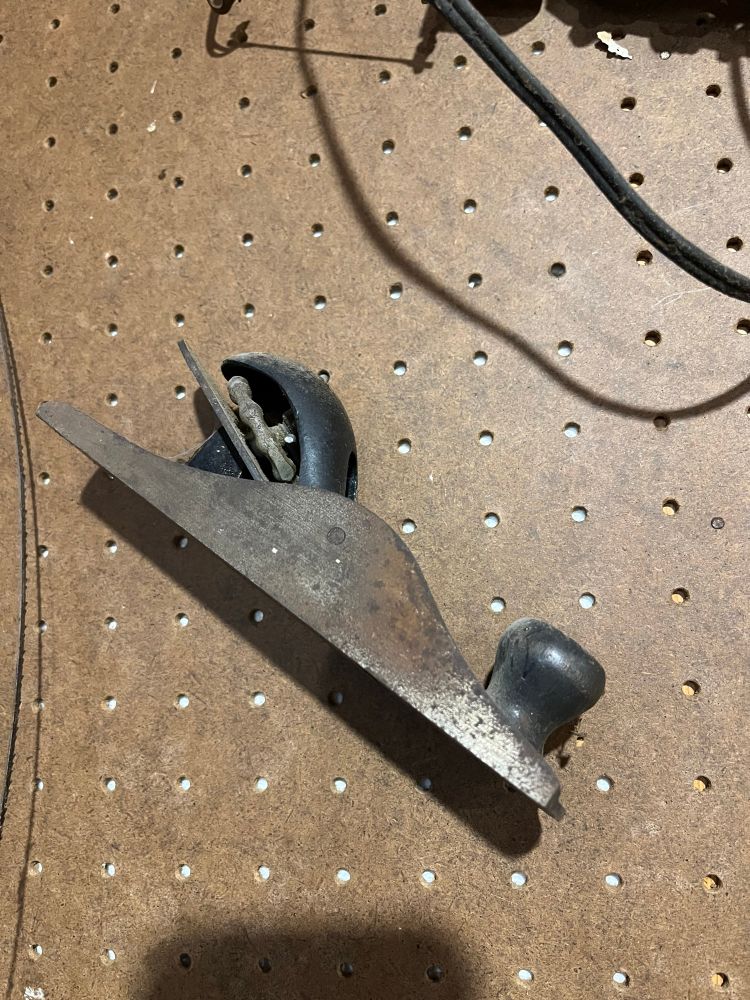 A small antique hand plane hanging on a workshop pegboard, rusty and dusty.