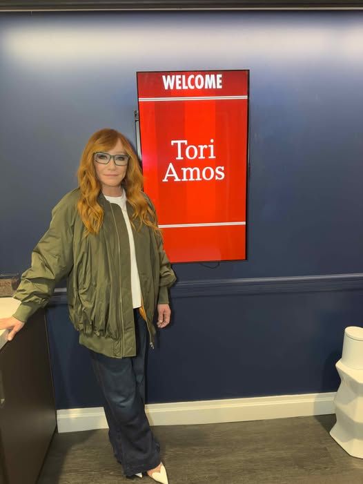 Tori Amos, a white woman with long red hair stands in front a red sign that says "Welcome Tori Amos" in a room off the set of the MSNBC television program Morning Joe. She is wearing a white shirt with an olive jacket, blue pants, and white heels.