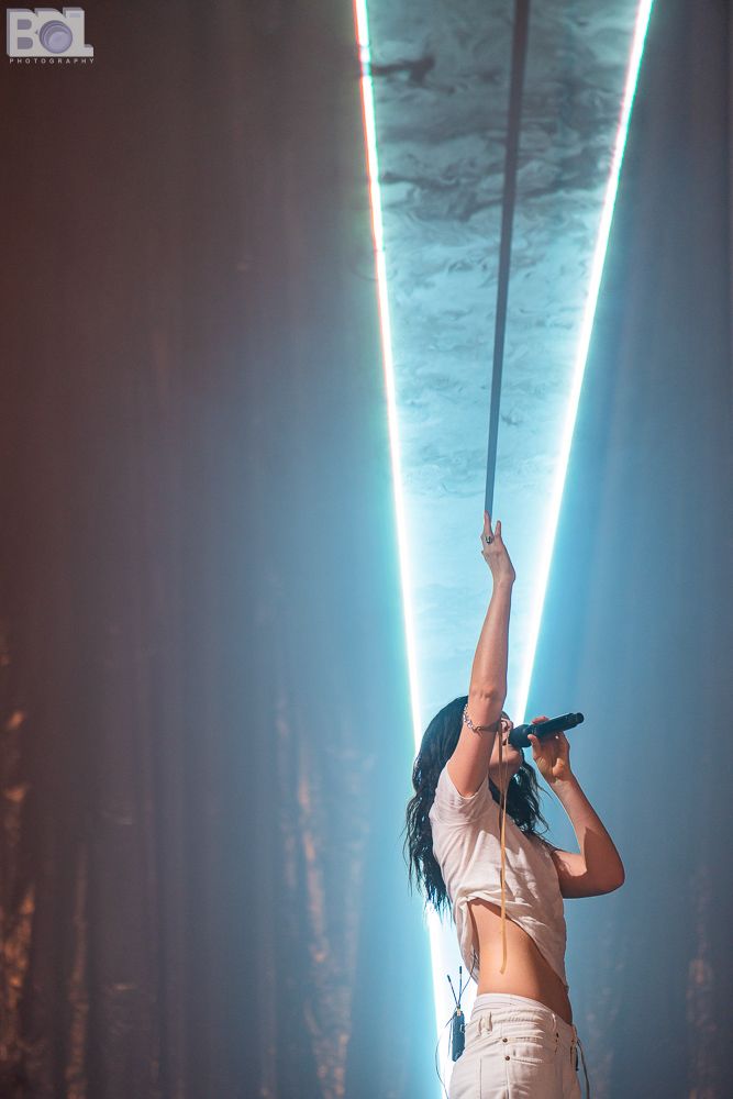 The singer Lorder is wearing white pants and a white shirt and singing with the microphone in her right hand. Her body is turned towards the right side of the photo and her left hand is reaching up to an aqua colored light and casting a shadow on it.