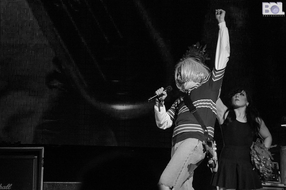 A black and white photo of Weird Al in an outfit that resembles Kurt Cobain. He has one arm raised in the air and holding the microphone with his other hand and is singing into it. There is a cheerleader behind him.