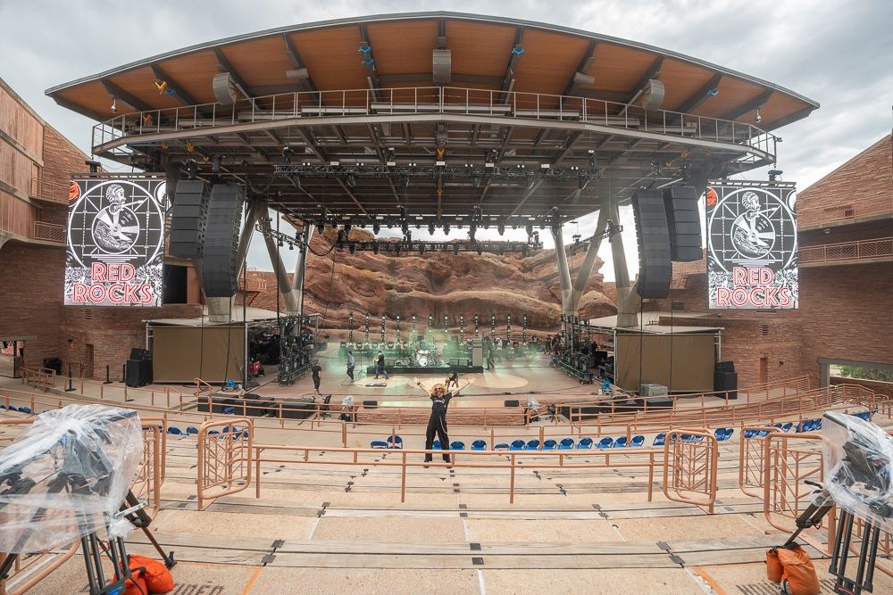 A man in black overalls has his hands in the air with the Red Rocks stage behind him.