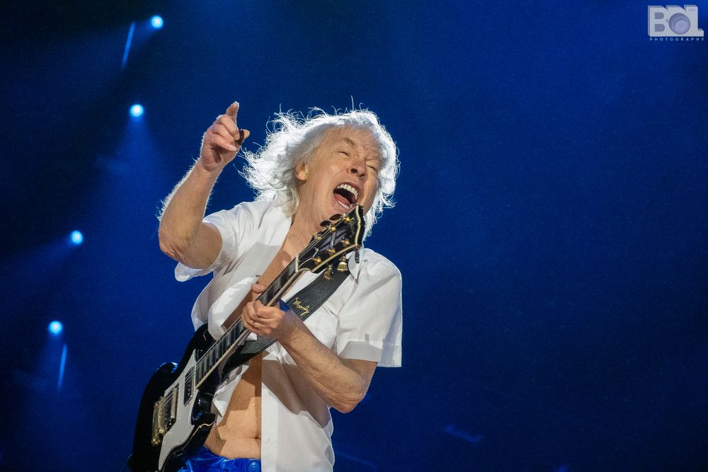 Angus Young of AC/DC in a white shirt and blue shorts pointing and yelling as he plays the guitar.