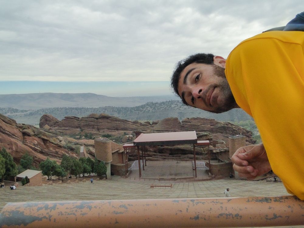 A man in a yellow shirt leans over a rail and looks directly at the camera with the venue Red Rocks in the background. His face has a 5 o'clock shadow and his black hair is short. 