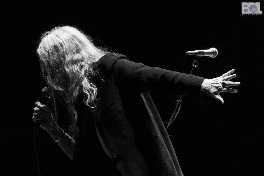 A black and white photo of Patti Smith singing into microphone wearing a black jacket. She is leaning down and to the left as her right hand holds the microphone and her left hand is extended behind her.
