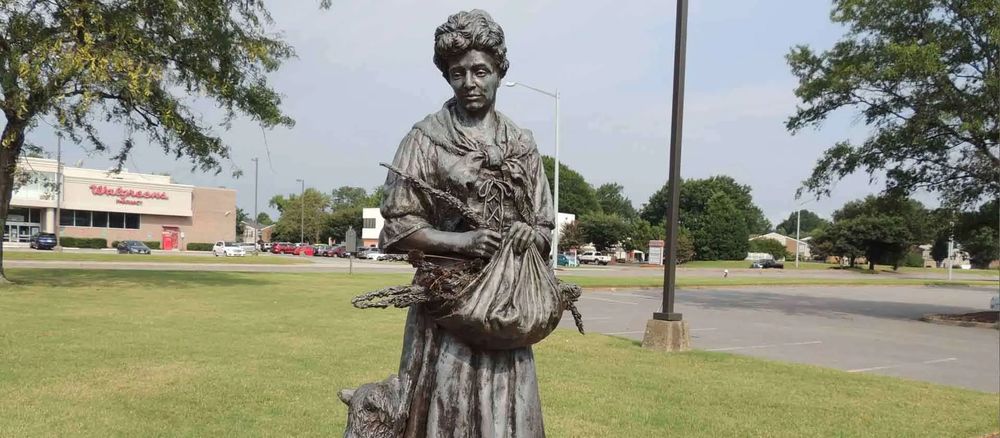 A picture of the Grace Sherwood Statue. She is depicted holding a basket of rosemary with a raccoon next to her.