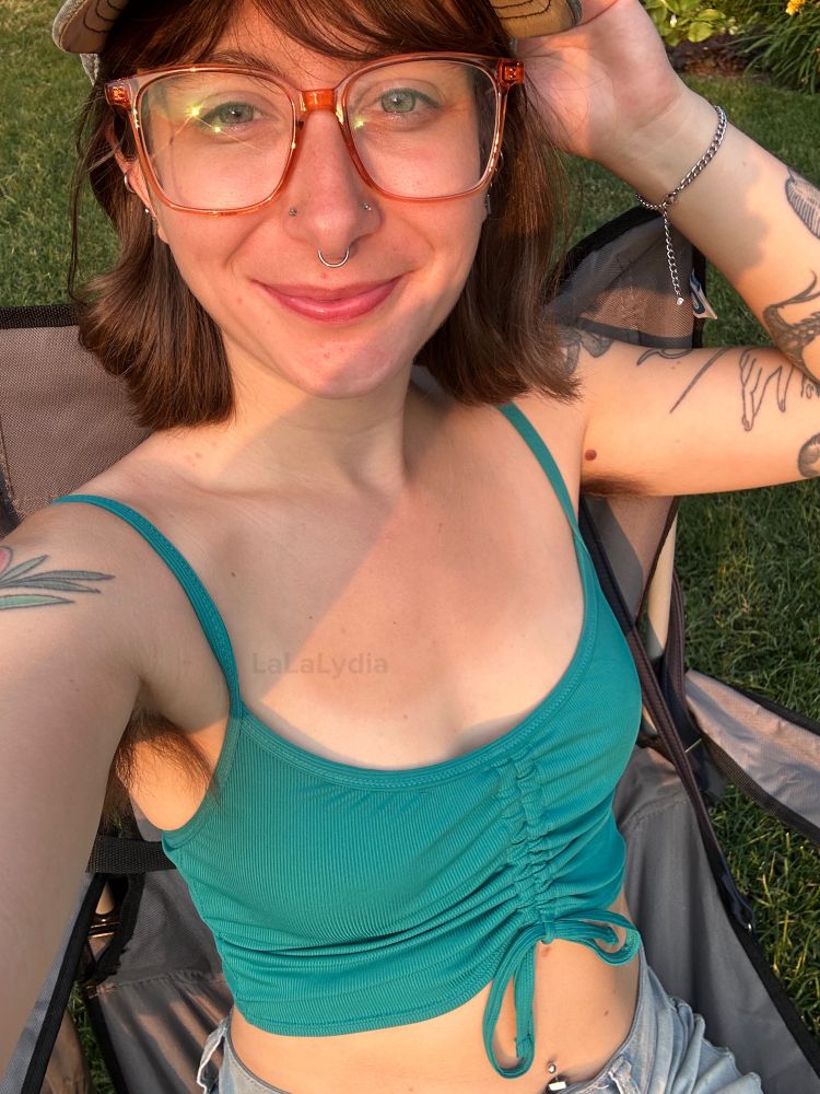 Lydia smiling, wearing jean shorts that show my bellybutton ring and tightly tied crop top that shows my arms and hairy armpits. I look tan and sit in a camping chair on a grassy lawn. I’m wearing big eye glasses, have nostril studs and a septum ring in and have short brown hair. 