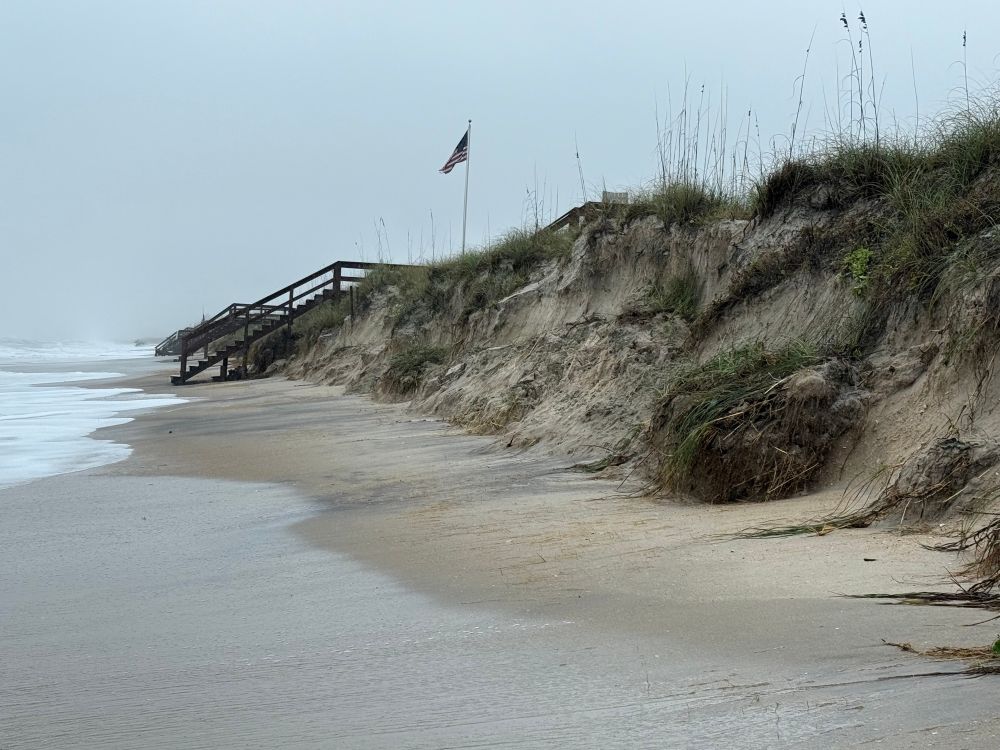 Dunes cut back like a cliff face, with the sea oats clinging to whatever they can.  Broken beach access stairs are visible farther down the beach 