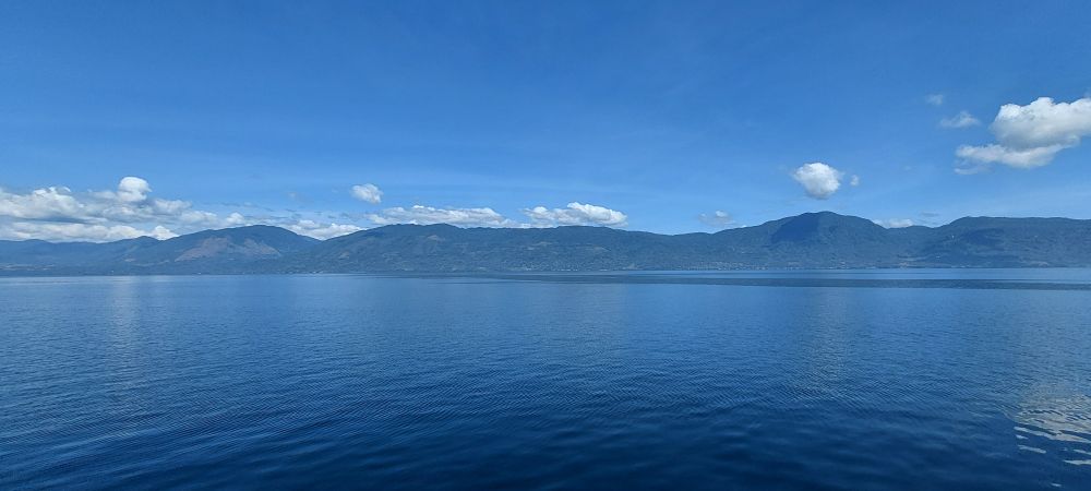 a view of a lake with a blue sky, white clouds hover over the lake and the mountains stand... not so tall, over on the horizon.