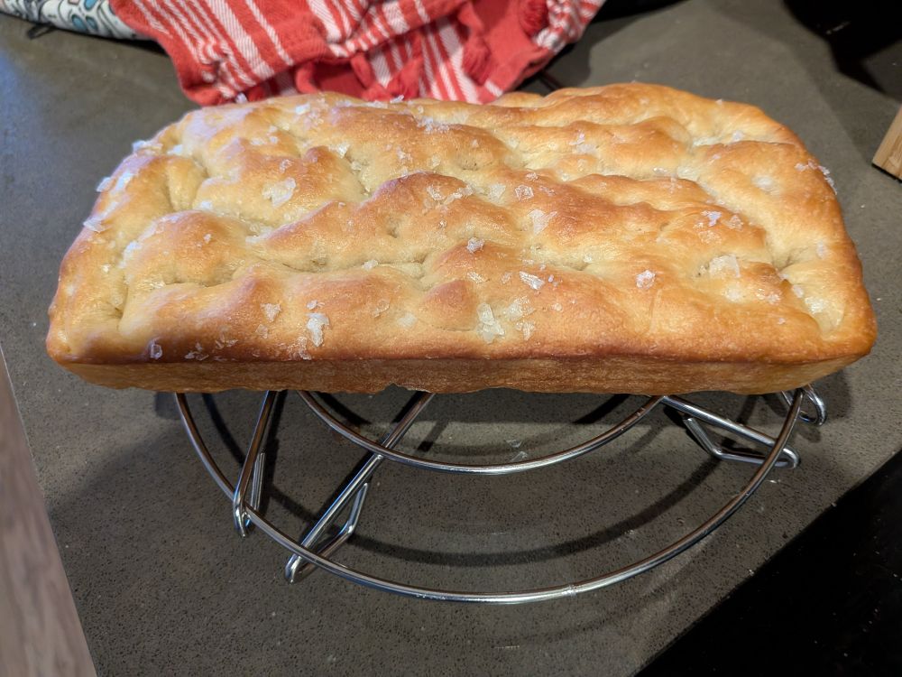 A small focaccia loaf with flaky salt on top. It's sitting on the trivet from my Instant Pot to cool. You can see a tea towel behind it on the counter.