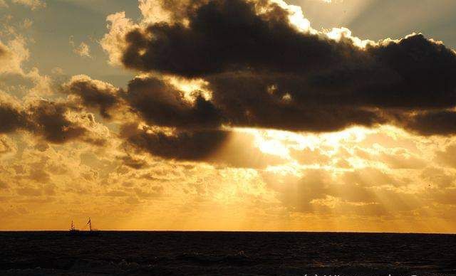 An orange coulored sunset with the sun hidden behind a cloud and the sun rays shining down to the sea. Just on the horizon a fishing boat is visible 