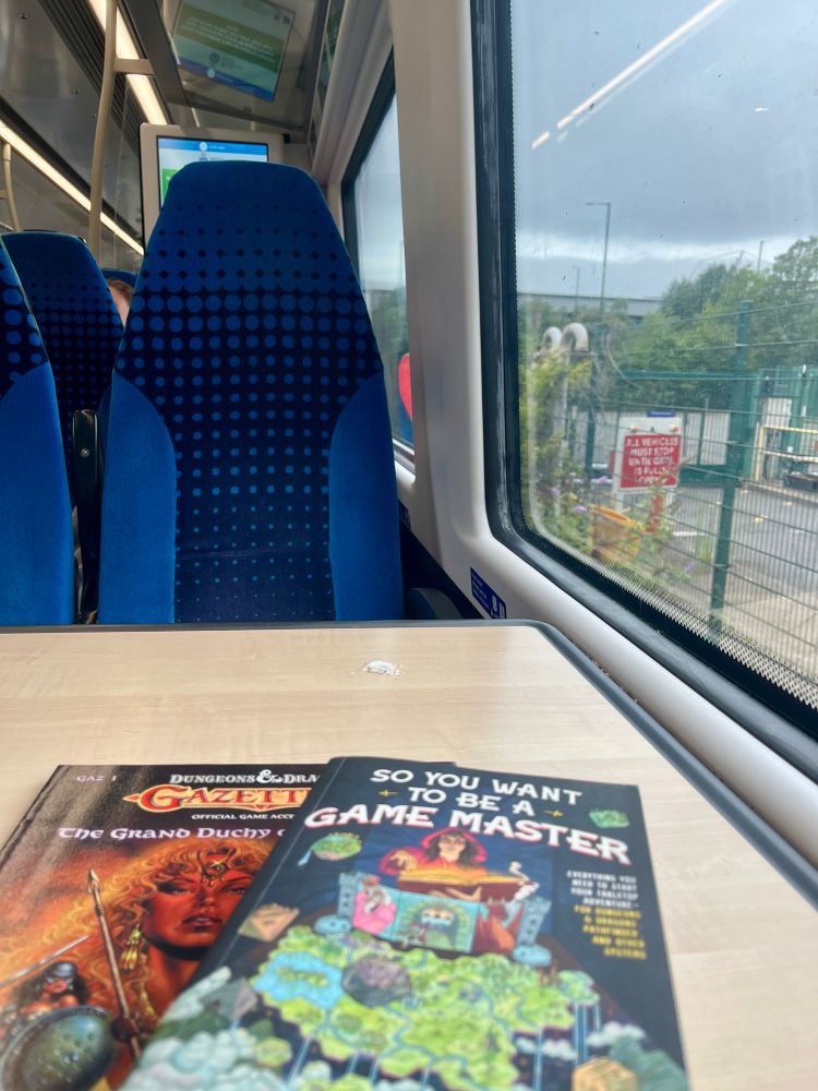 Photo of a window seat in the Northern train to Leeds via Sheffield. A copy of the GAZ1 Karameikos book sits on the table underneath a copy of “So You Want To Be A Game Master”. The view from the window is urban Nottingham.