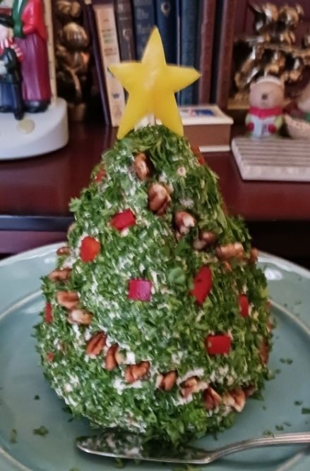 A cheese ball decorated to look like a Christmas tree. It is covered in parsley with red pepper ornaments, a pecan garland, and a yellow bell pepper star. It's on a teal plate with a butter knife.