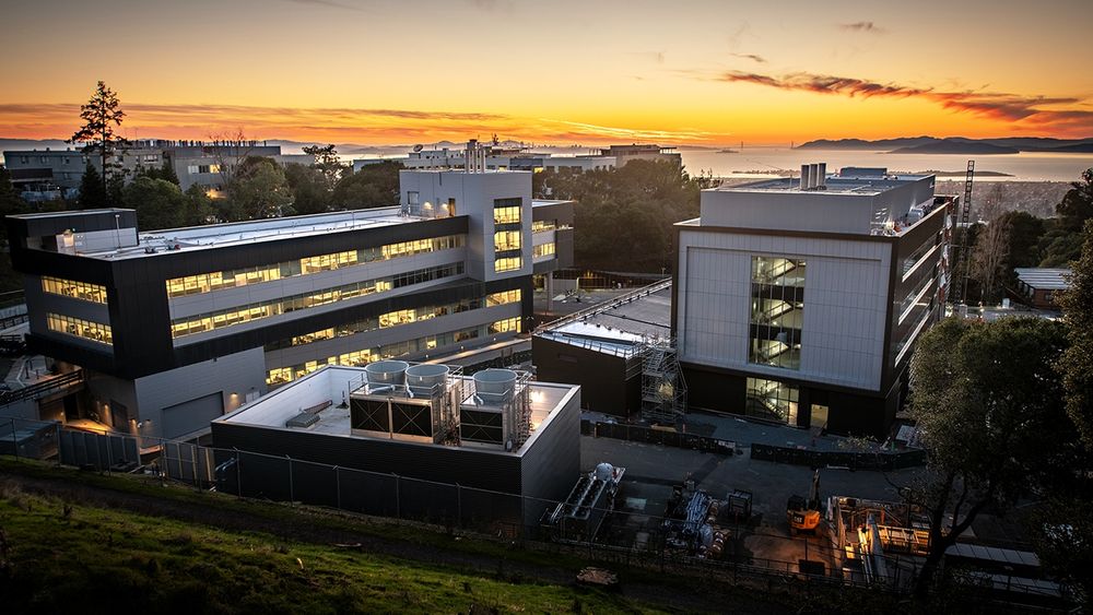 Buildings on the Lawrence Berkeley National Lab campus, with the San Francisco Bay and the Golden Gate Bridge in the background. It's sunset time, the sky is orange.  On the left is the Integrative Genomics Building (IGB), which houses the JGI, NMDC and KBase. On the right is the BioEPIC building.