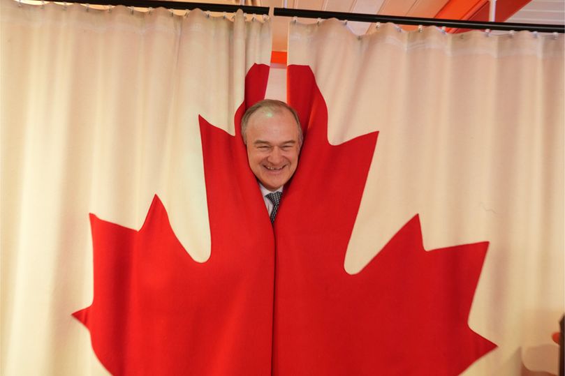  Ed Davey peeks through a Canadian flag curtain, his face framed by the maple leaf, smiling.