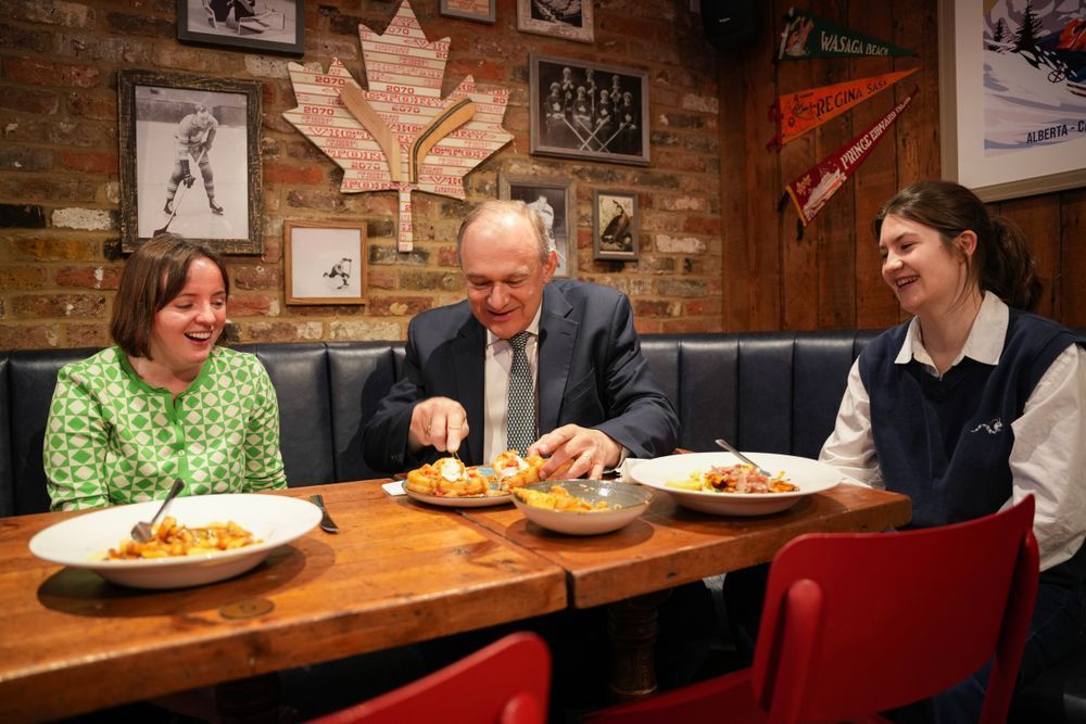 Ed Davey shares a meal with two women in a Canadian-themed pub, enjoying poutine and laughter. Hockey memorabilia and a maple leaf made of hockey sticks decorate the background.
