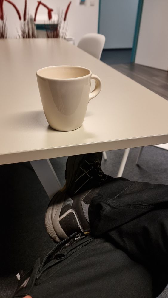 Man sieht einen Beigefarbenen Tisch, ein Bein angewinkelt auf dem anderen ruhend und eine Tasse Kaffee.