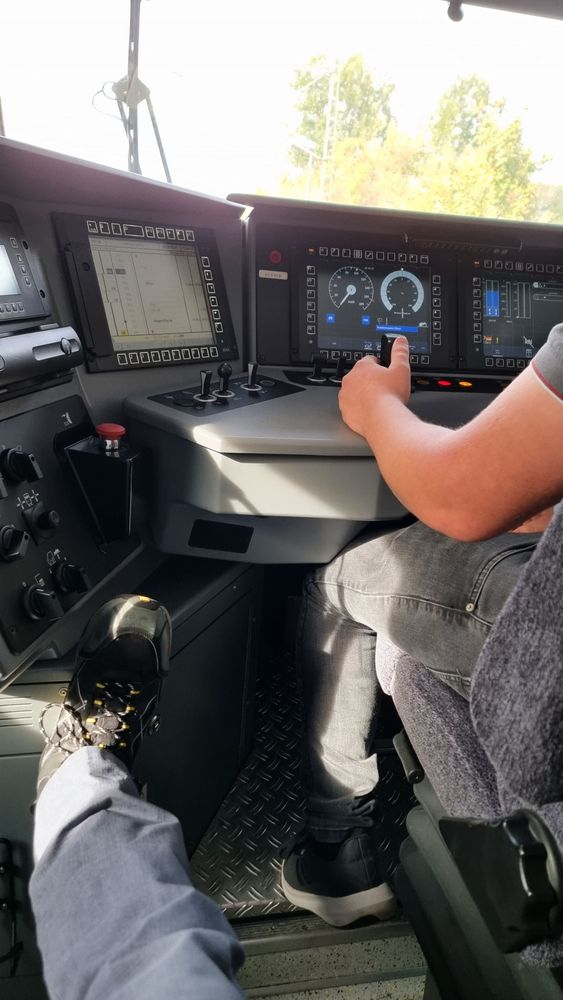 Blick auf einen Führerstand eines Flirt 3 Zuges. Azubi hält gerade für einen Fahrgastwechsel an. Man sieht den elektronischen Buchfahrplan, den Tacho und sämtliche Hebel wie die Wachsamkeitstaste, den Fahr-Brems Hebel und die Knöpfe für die Türen.