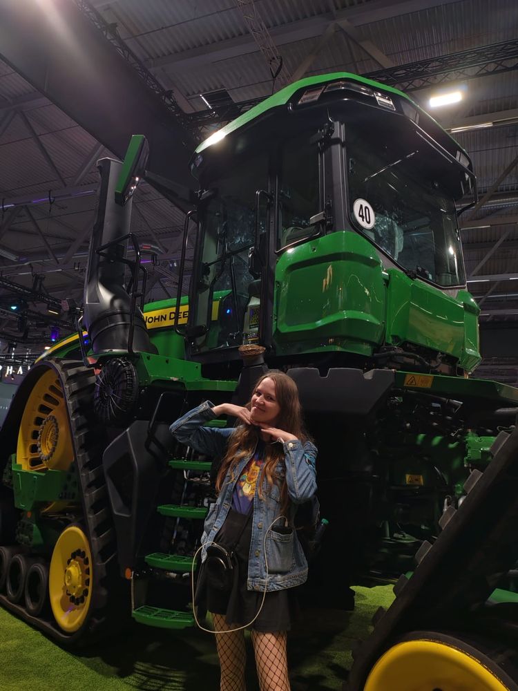 Eine junge Frau posiert lächelnd vor einem riesigen grünen John-Deere-Traktor auf einer Messe. Sie trägt ein schwarzes Outfit mit Netzstrumpfhose und Jeansjacke und legt die Hände verspielt unter ihr Kinn.