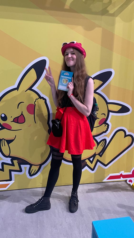Eine junge Frau posiert vor einer großen Pikachu-Wand, trägt ein rotes Kleid mit schwarzem Oberteil und Hut und hält stolz ihren Pokémon-Trainerpass hoch, während sie ein Peace-Zeichen macht.