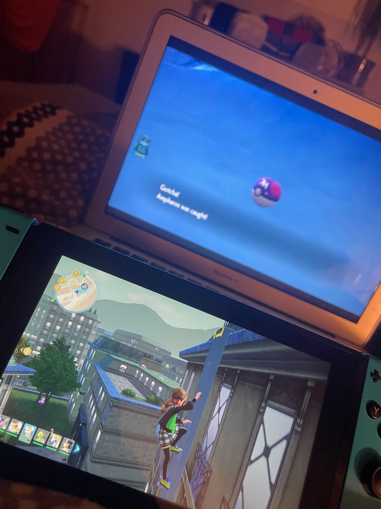 A cozy evening setup showing two screens: on a laptop in the background, a Master Ball is used to catch an Ampharos in Pokémon Scarlet or Violet, while on a Nintendo Switch in the foreground, a female character climbs a tall building in Pokémon Legends: Z-A. The scene is softly lit and feels relaxed and nostalgic.
