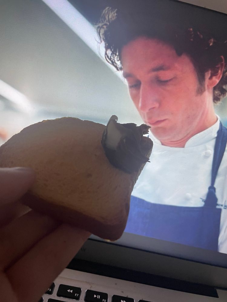 A hand holds a piece of rusk with a spoonful of chocolate spread in front of a laptop screen. On the screen, a scene from The Bear shows chef Carmy looking down with a serious expression.