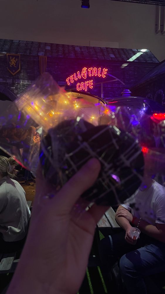 Ein Handfoto mit einem verpackten Cookie im Vordergrund, unscharf gehalten. Dahinter eine Szene mit Lichterketten und einem leuchtenden Neon-Schild mit der Aufschrift „Telltale Café“.