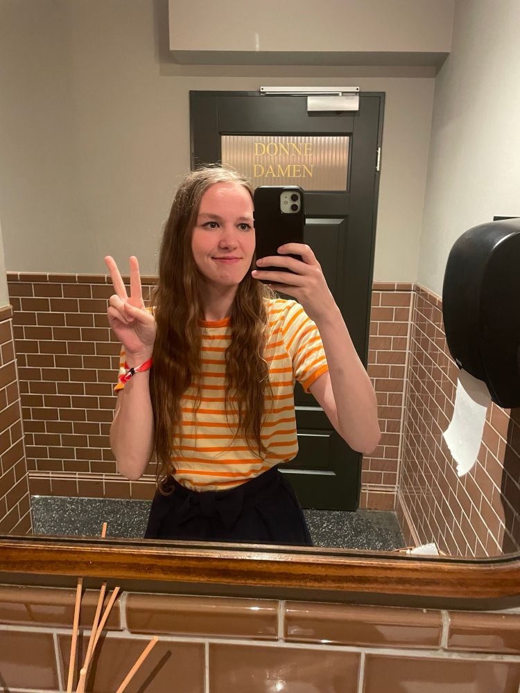 Eine Person macht ein Spiegel-Selfie in einer Toilette mit braunen Fliesen. Sie trägt ein orange-weiß gestreiftes T-Shirt und einen dunklen Rock, hält das Handy hoch und zeigt mit der freien Hand ein Peace-Zeichen.