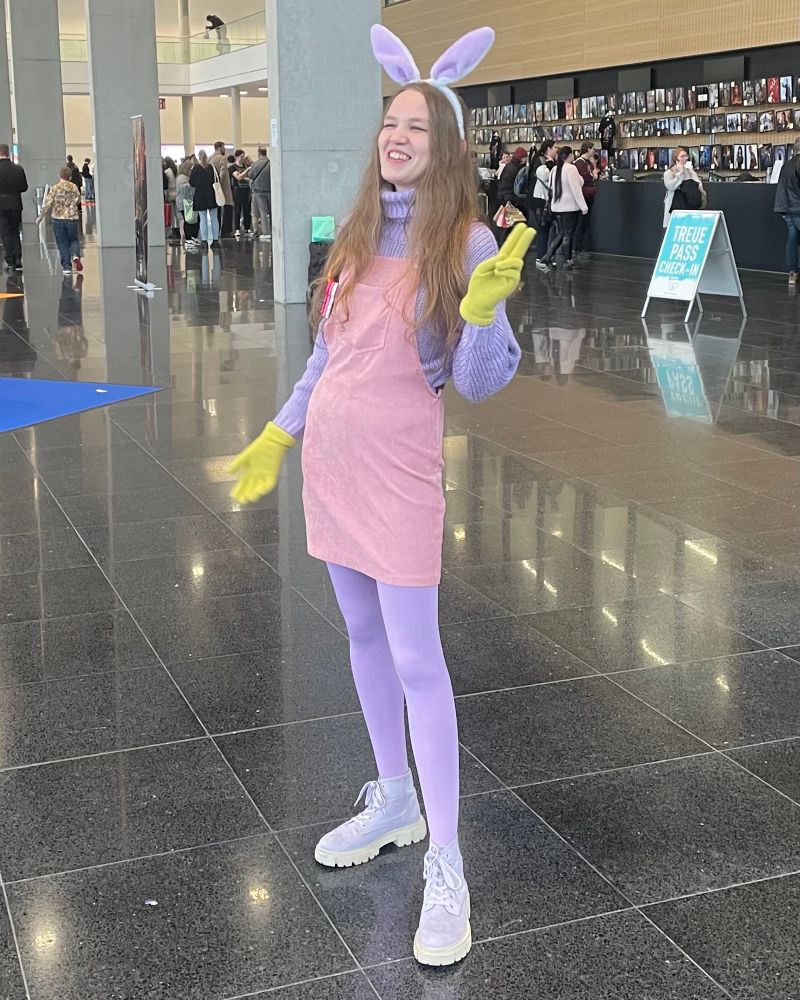 A young woman in a pastel bunny-inspired Jax cosplay stands laughing in a convention hall. She wears lavender bunny ears, a purple turtleneck sweater, a pink dungarees dress, lilac tights, chunky light-purple boots, and yellow gloves. She poses with one hand raised in a playful peace sign.