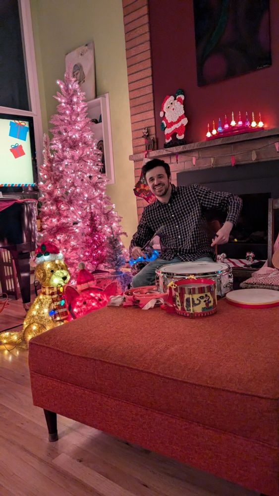 Alex at a Christmas-decorated house, smiling, holding drum brushes, about to play a snare drum