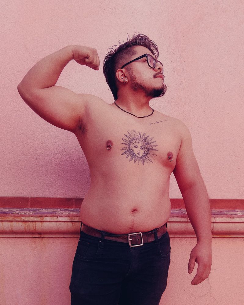 Foto de medio cuerpo de Obed. De pie, flexiona el brazo para mostrar el bicep. El otro brazo cae a un lado, mira a su lado izquierdo, mostrando el lado de la cara. Obed es de piel clara, cabello corto y negro, barba y bigote pequeños. Nariz chata, pequeña, labios delgados y rosas. Tiene el cabello muy corto a los lados, largo de arriba. Tiene un tatuaje de sol en el medio del pecho y una frase en la clavicula. Usa pantalón negro, cinturón café y lentes de pasta amplios, negros y múltiples aretes plateados en su oreja y argollas plateadas y gruesas en los pezones. Tiene un collar delgado y corto negro. Al fondo una pared color crema y un pequeño borde con diseño pecho de paloma a la altura de la cintura.