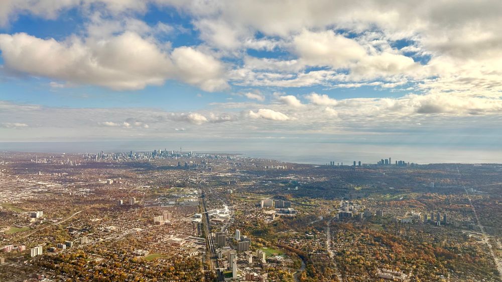 A view of Toronto in early November from an airplane recently departed from Pearson, facing downtown from around Lawrence and Weston. Scattered clouds cast mottled shadows. The million trees are on average a rust colour. 