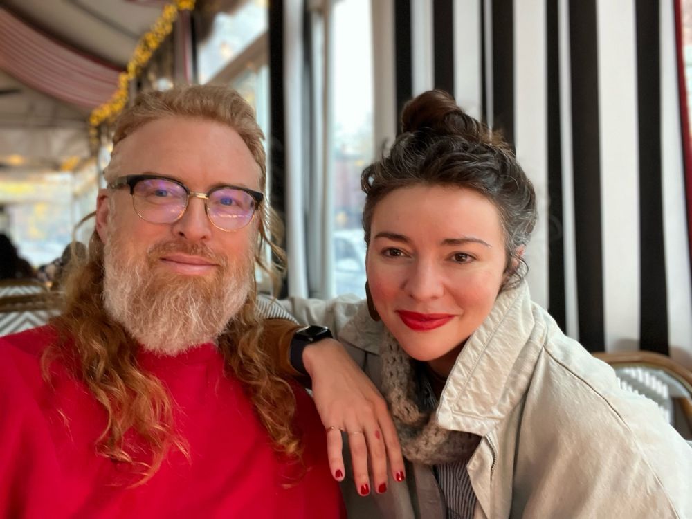A caucasian male-presenting person with a full grey beard and a long, curly red mullet wears a red, crew-neck sweatshirt and fills the left half of the frame. Filling the right frame, is a female-presenting, caucasian woman wearing her dark brown and gray hair in a bun, a light gray denim jacket with the collar popped. She leans in to the man and her right hand, nails painted red, lingers on his left shoulder. She wears red lipstick. They are both smiling. 