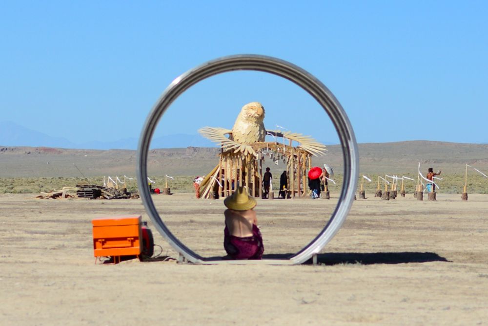 A woman sitting in a large tube for shade as she gazes at a large, wooden eagle statue in the distance. 