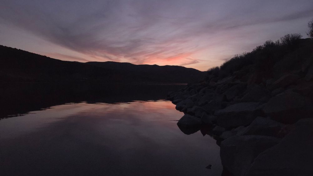 A sunset with light pinks and pale blues. There are some whispy clouds that range from gray to pink in color. The silhouette of a mountain range is in the distance. Both the sunset and mountain range are reflected in the foreground by a lake with still water.