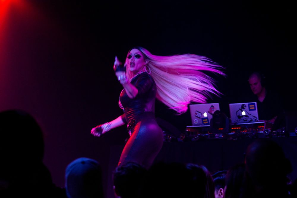 Image shows drag performer Morgan McMichaels during a performance. She is mid dance move and her long blonde hair has swung behind her, which catches the pink color from the stage lights.
