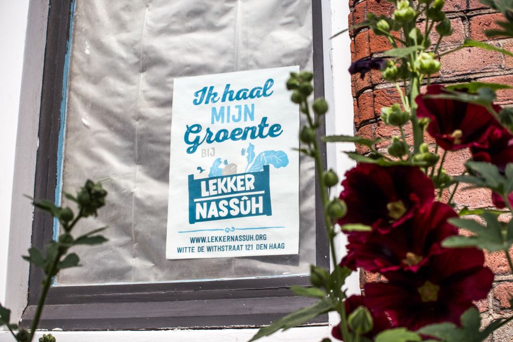 Een poster in een raam met in blauwe tekst ‘Ik haal mijn groente bij Lekkernassûh, www.lekkernassuh.org, Witte de Withstraat 121 Den Haag’. Op de voorgrond bloeit een donkerrode klaproos.