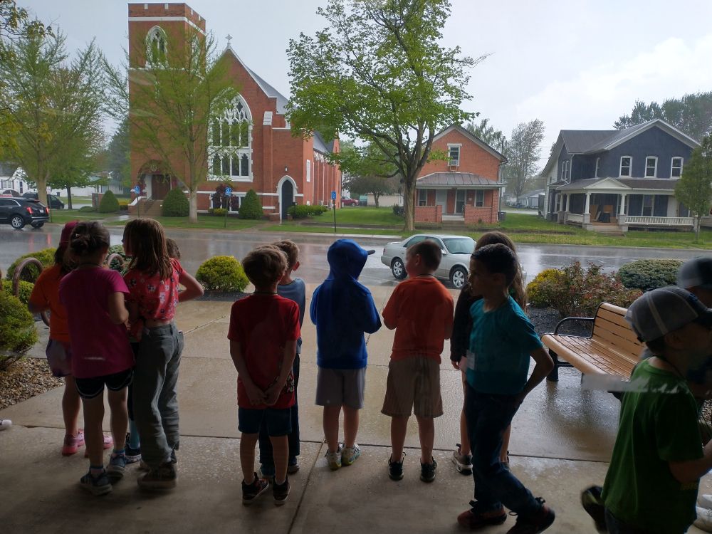 Kids standing on a sidewalk underneath an overhang, watching as it rains. 