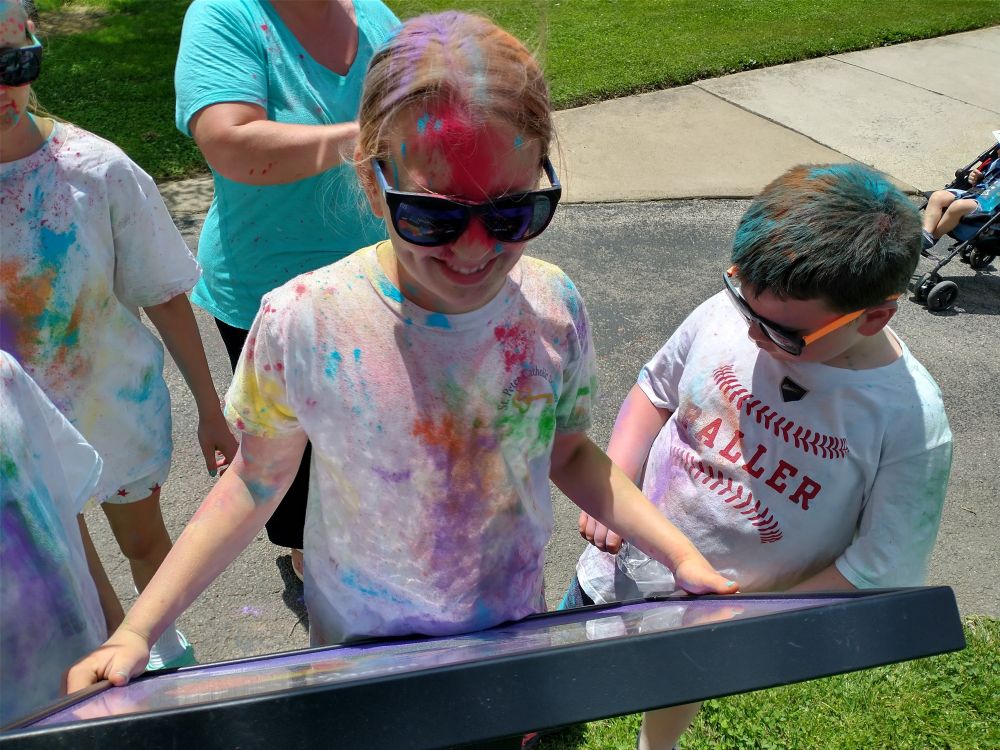 A group of kids covered in colored powder standing at a podium and reading a page of a book. 