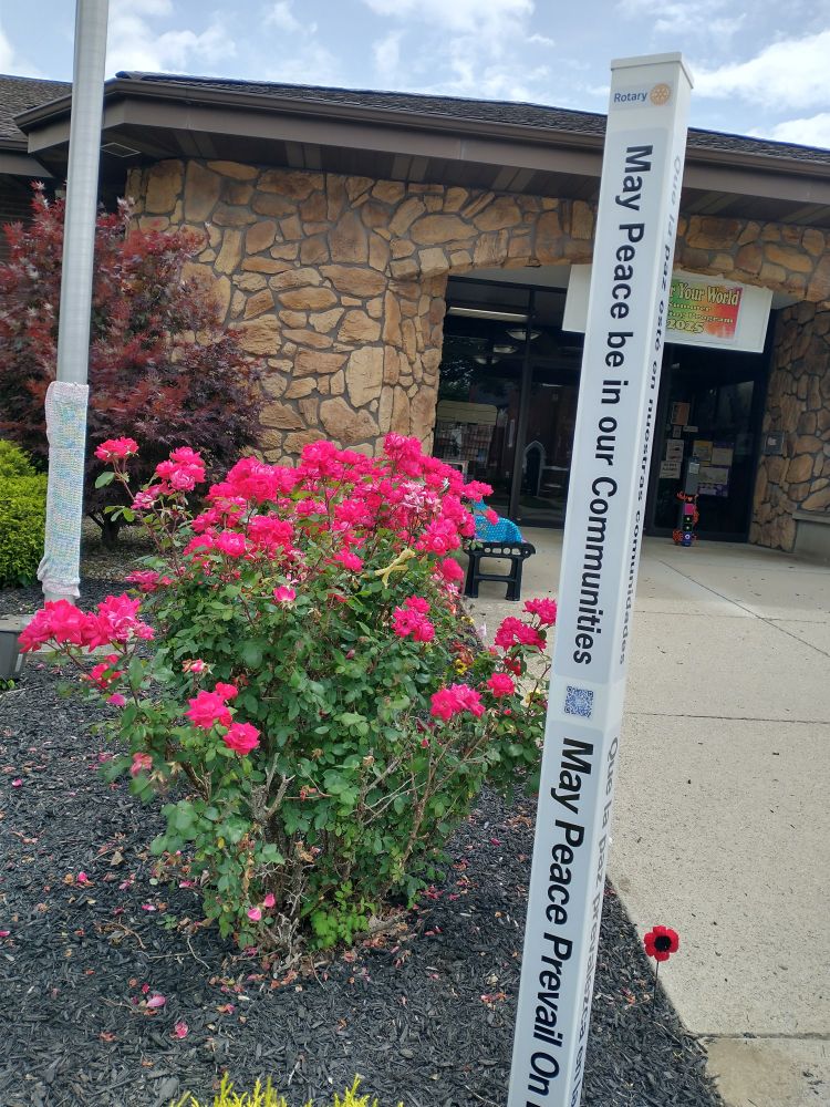 A pole in a rose garden. The pole has "may peace be in our communities. may peace prevail on Earth" on all sides in different languages. 