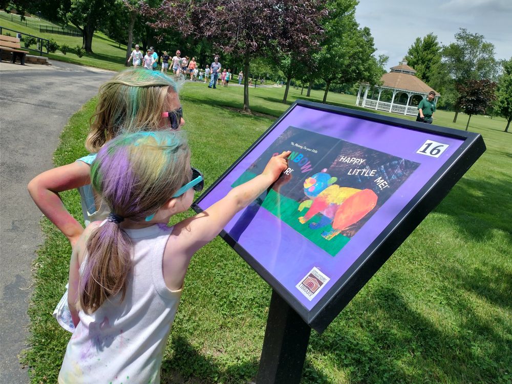 Two little girls covered in colored powder are reading a book page on an outside podium at a park. 
