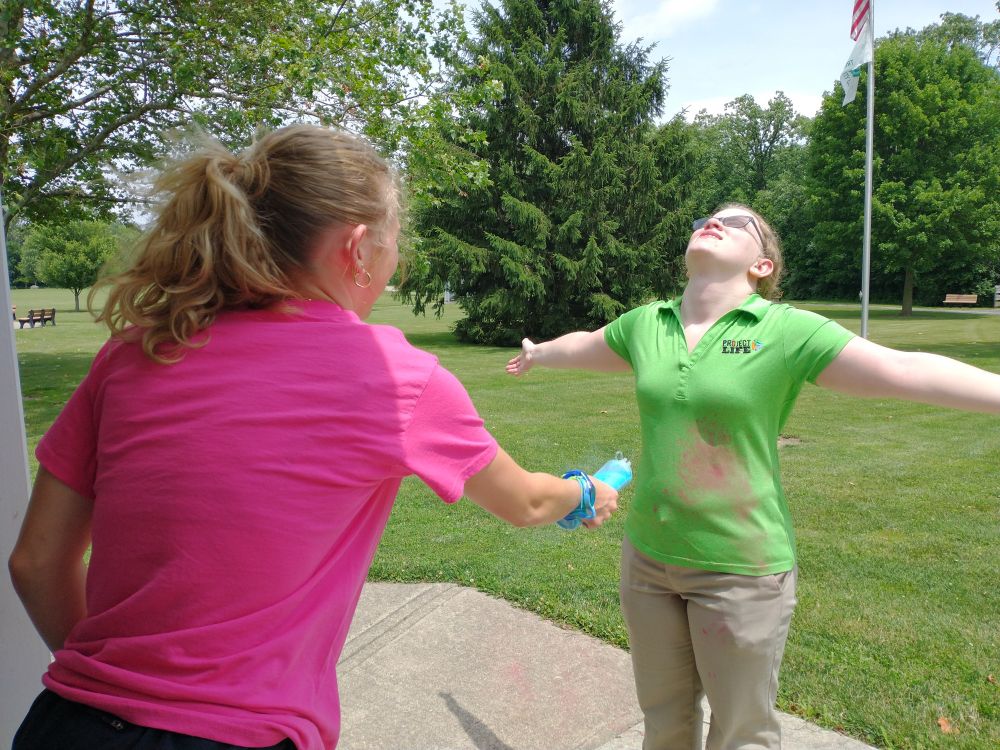 One woman squirting colored powder on another at a park. 