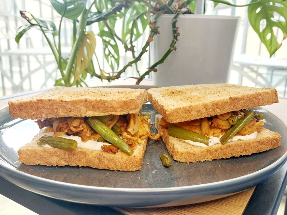 two wholegrain sandwiches filled with fried chunks, sour cream and green beans on a dark grey plate; Monstera in the background
