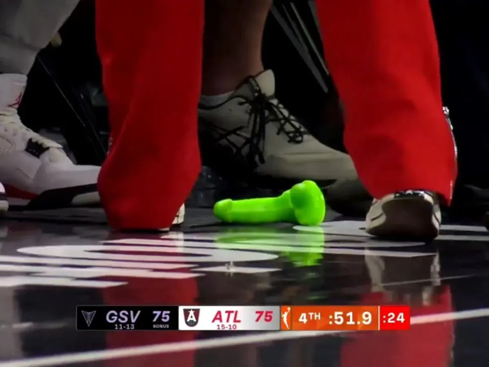 Imagem mostra um dildo verde no chão de uma quadra de basquete, há pernas de várias pessoas em volta.