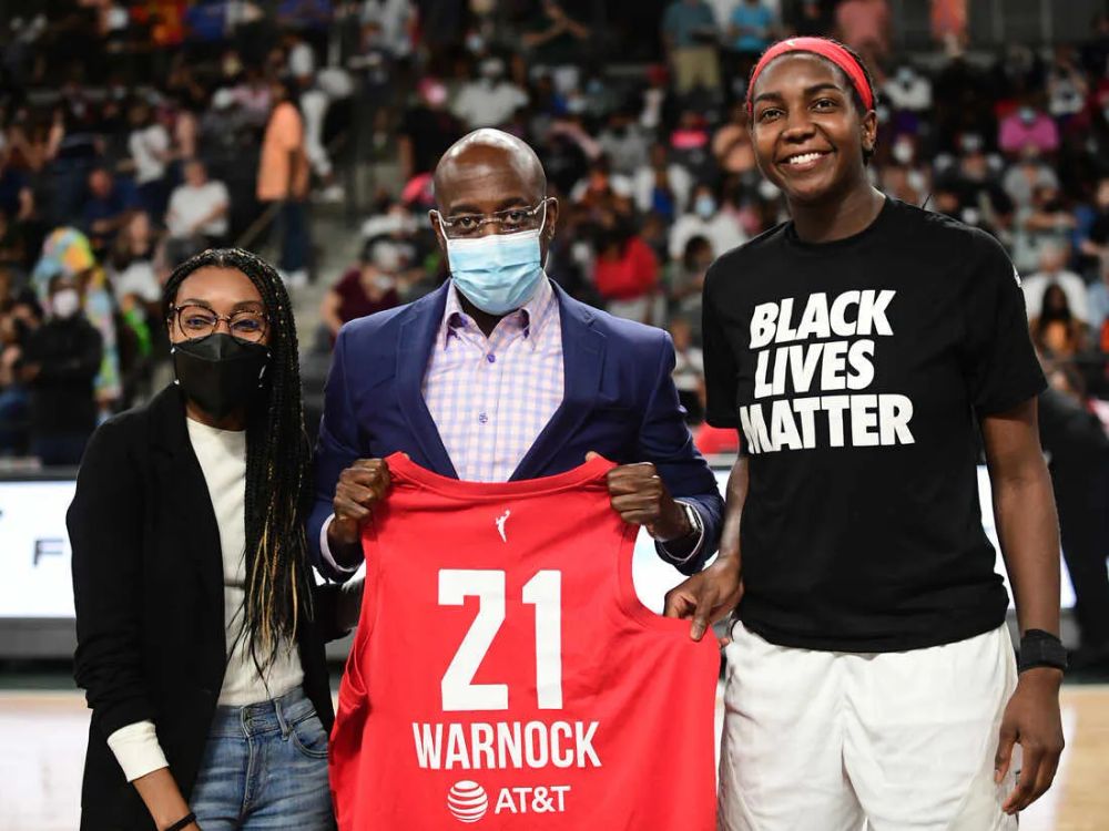Imagem mostra uma jogadora da WNBA com uma camisa com os dizeres "Black lives matter", há um homem segurando uma camisa vermelha e uma mulher de máscara ao lado dela.