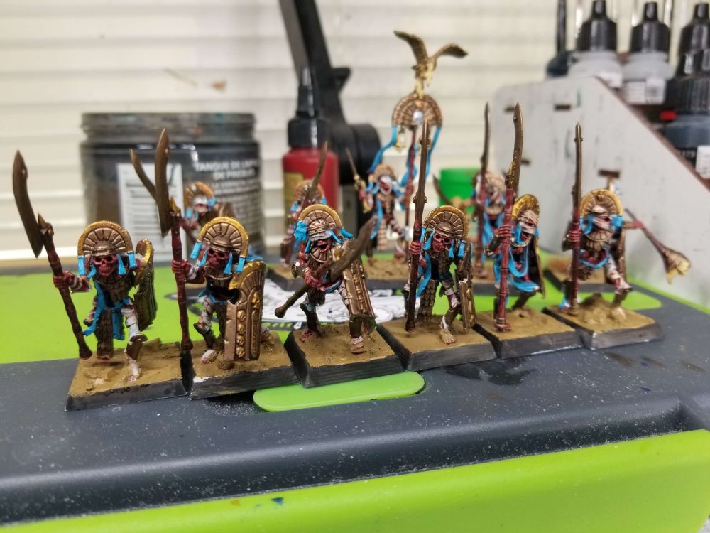 6 tomb guard arranged in a row with halberds. Each one has a different pose with dirty brown bases and bronze shields and crowns. They have blue for their clothing and highlight colors, though have dirty bandages. 
