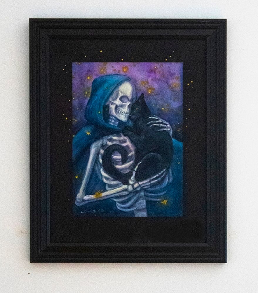 Boundless, framed. Shows skeleton snuggling black cat, surrounded by fireflies with a black frame and black embellished mat.
