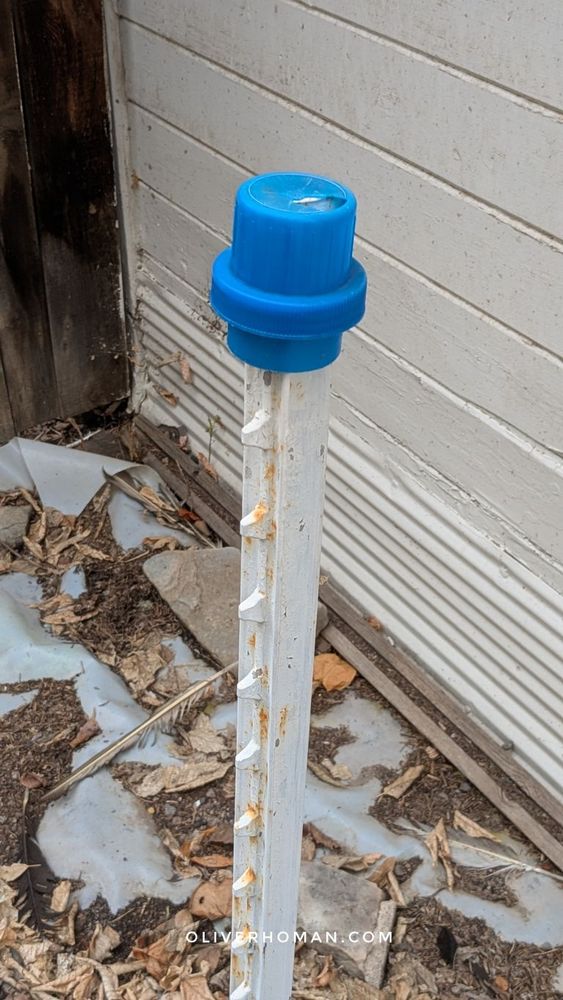 A blue laundry detergent bottle cap on top of a metal stake in an alleyway. Fall leaves line the dirt alley next to a fence and white shack.